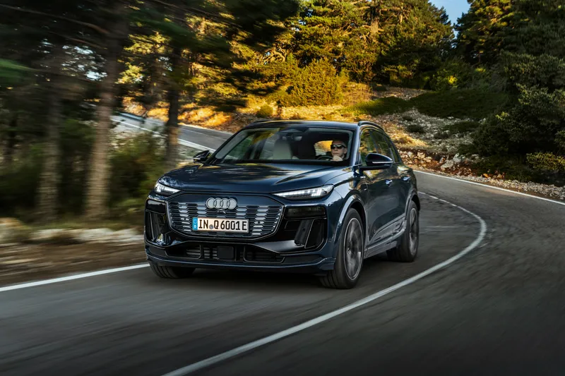 Audi Q6 e-tron azul oscuro en curva de carretera de montaña entre pinos al atardecer